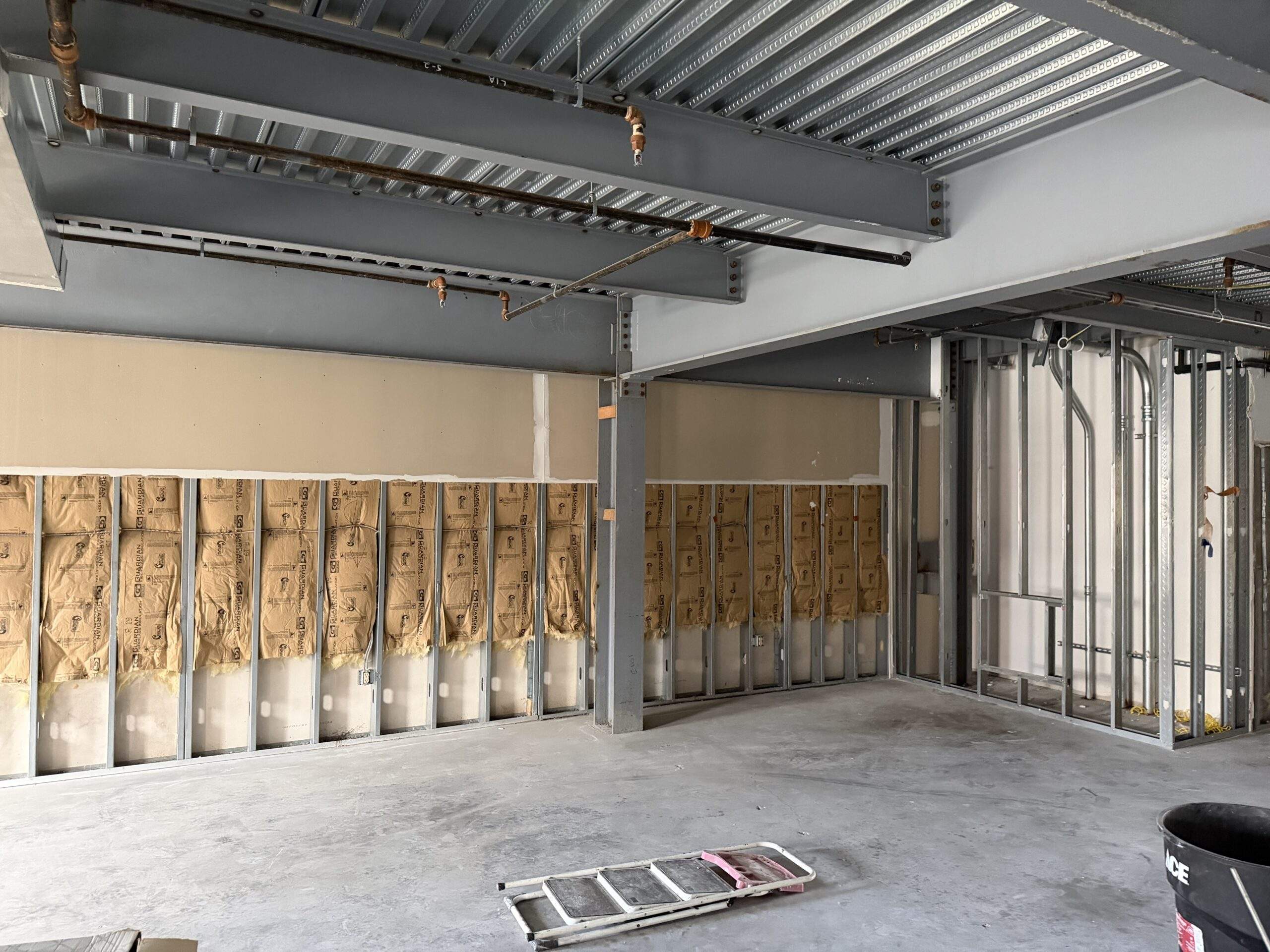 Interior view of unfinished commercial space with exposed metal studs, insulation, and ceiling infrastructure.